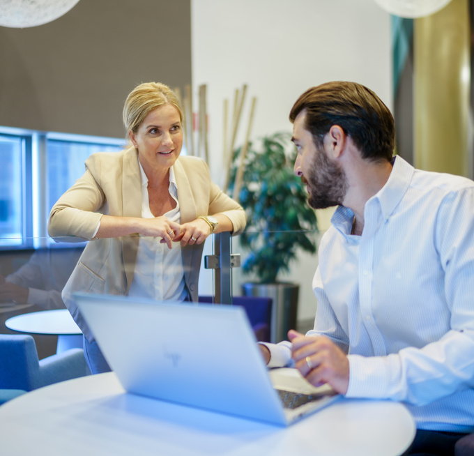 Een medewerker leunt op iets en praat met een andere medewerker die achter zijn bureau zit