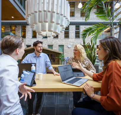 Vier medewerkers zitten aan een hoge tafel achter hun laptop en zijn aan het overleggen