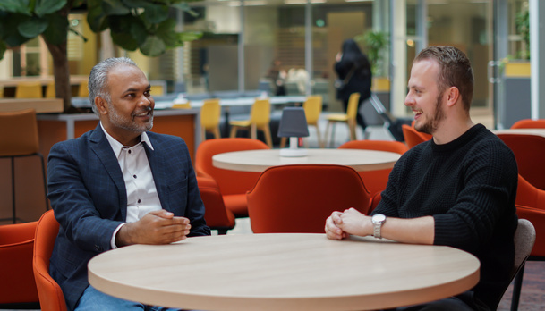Twee medewerkers zitten in de hal aan een tafel en kletsen