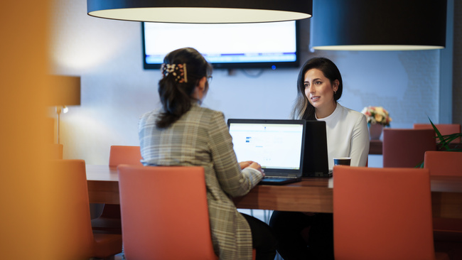 Twee medewerkers die in gesprek zijn en zitten aan een bureau