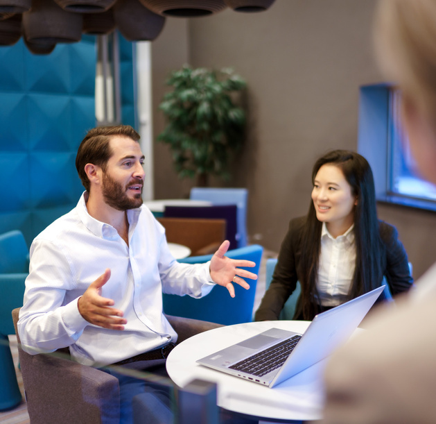 Een medewerker zit aan een tafel met zijn laptop en vertelt iets aan een andere medewerker